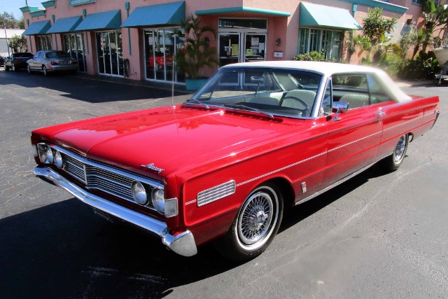 Import American cars from USA image of a red 1966 Mercury S-55 parked in front of a pink fronted store on asphalt.