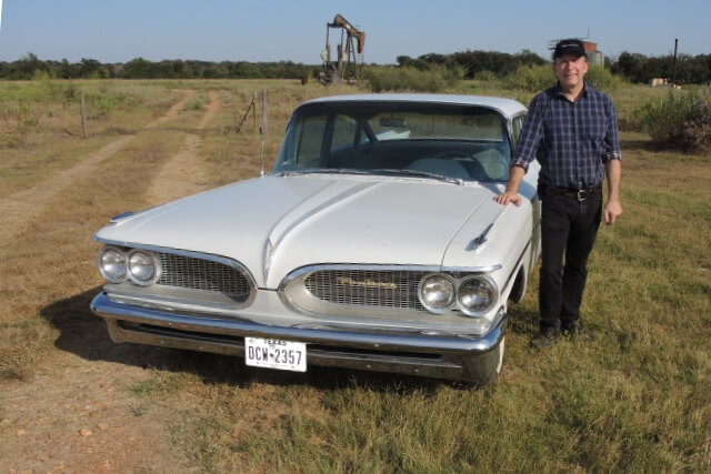 American white car parked in a field with an oil pump in the backgrond with the inspector next to the vehicle.
