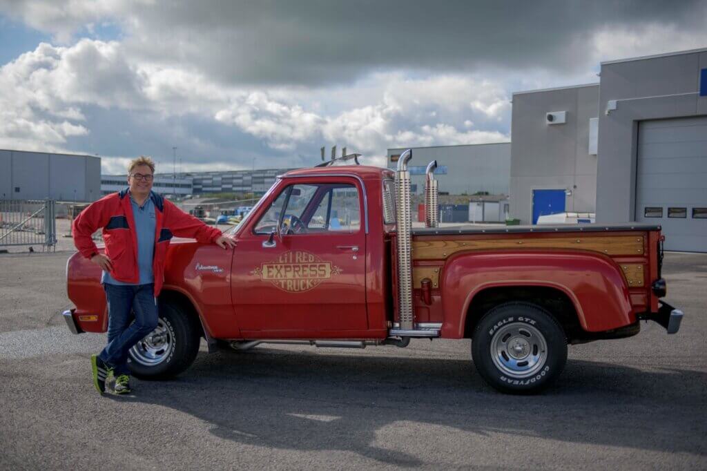 Door2Door imported a red 1979 Dodge D150 Lilttle Red Express to Sundbyberg for a satisfied customer standing next to the car with his hand on the engine head in an industrial area